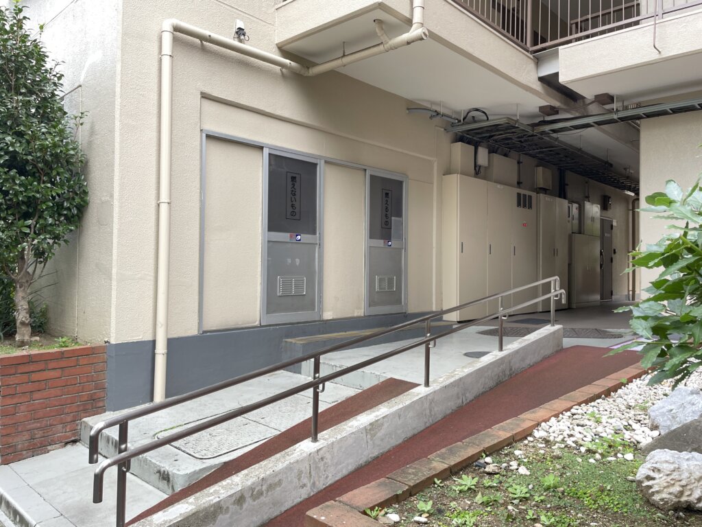 マリンシティダイヤモンドパレス敷地内のスロープとゴミ置き場。手すり付きの緩やかなスロープがあり、その奥に「燃えないもの」などの表示があるゴミ収集スペースが設置されている。