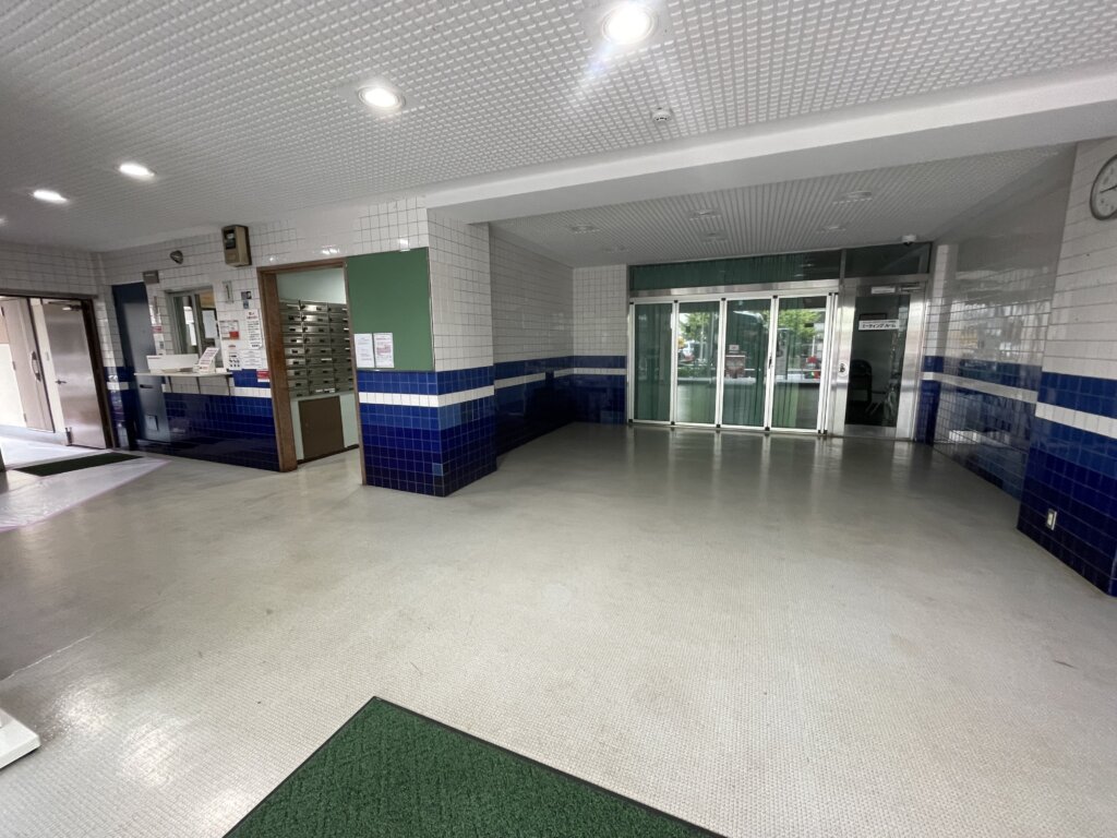 マリンシティダイヤモンドパレスのエントランスホール内部。白と青のタイル張りの壁で、左側に集合ポストと管理人窓口があり、奥にはエレベーターホールへの自動ドアが見える。
